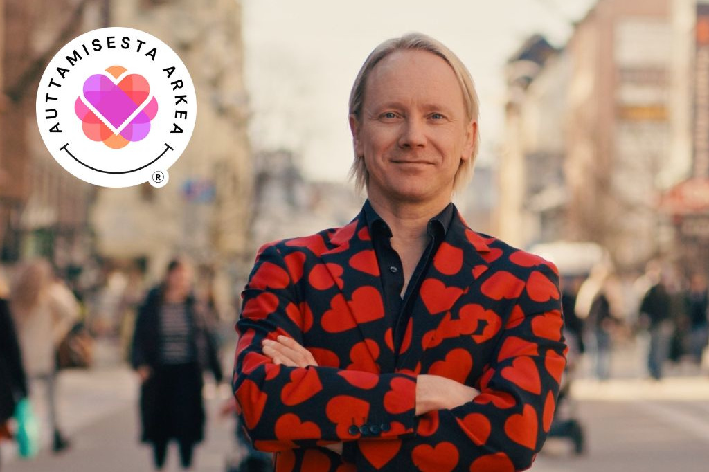 Olkapäille ulottuvilla vaaleilla hiuksilla varustettu mies hymyilee kameralle kädet ristissä. Hän seisoo sumealla kaupungin kadulla ja hänellä on yllään näyttävä musta bleiseri, joka on peitetty suurilla punaisilla sydämillä, mustan paidan päällä. Vasemmassa yläkulmassa on valkoinen pyöreä logo, jossa on teksti "AUTTAMISESTA ARKEA" värikkään, pikselöidyn sydänkuvakkeen ympärillä.