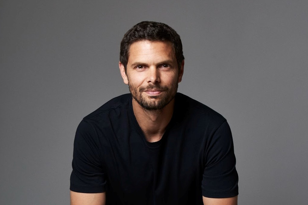 Studiomuotokuva keski-ikäisestä miehestä, jolla on lyhyet, tummat hiukset ja parta. Hänellä on yllään musta pyöreäkauluksinen t-paita, ja hän katsoo suoraan kameraan kevyesti hymyillen tasaisen harmaata taustaa vasten.