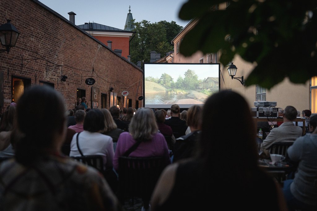Laaja, tunnelmallinen kuva yleisön takaa ulkoilmaelokuvanäytöksessä hämärän aikaan. Yleisö istuu eurooppalaistyylisellä sisäpihalla pitkän punatiilisen rakennuksen vasemmalla ja vaaleamman rakennuksen oikealla puolella. He katsovat elokuvaa, joka heijastetaan suurelle valkokankaalle, jossa näkyy maalaismainen maisema, joki ja puutalo. Taustalla rakennusten välissä näkyy vihreä kirkontorni iltahämärää vasten. Etualan yleisö on enimmäkseen siluettina.
