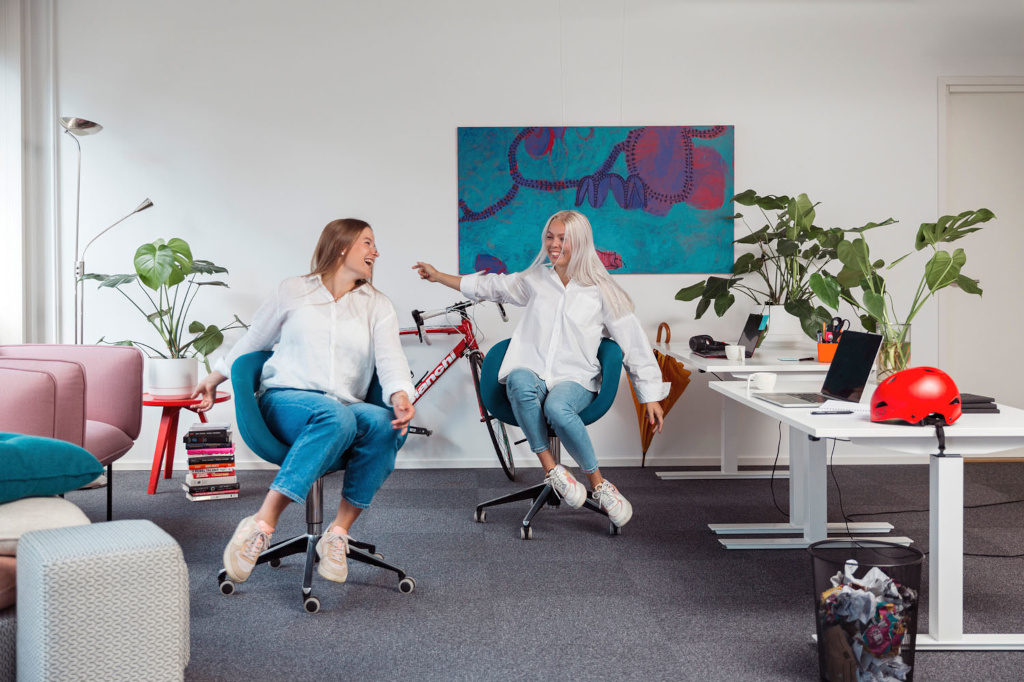 Laajakuva valoisasta ja modernista toimistotilasta, jossa kaksi nuorta naista pitää hauskaa. He ovat molemmat pukeutuneet valkoisiin kauluspaitoihin ja farkkuihin ja istuvat sinivihreissä pyörällisissä toimistotuoleissa nauraen yhdessä. Oikealla oleva nainen, jolla on pitkät vaaleat hiukset, osoittaa vasemmalla olevaa naista. Toimisto on kalustettu valkoisella seisomapöydällä, jolla on kannettavia tietokoneita ja punainen pyöräilykypärä, seinää vasten nojaavalla punaisella polkupyörällä suuren abstraktin maalauksen alla, useilla suurilla ruukkukasveilla ja rento oleskelualue, jossa on vaaleanpunainen nojatuoli etualalla.
